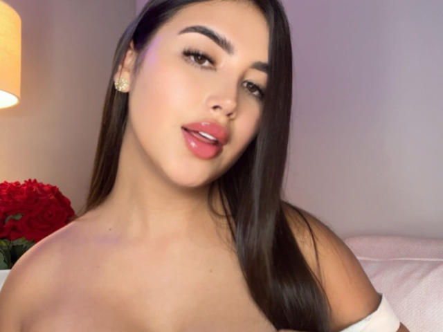 AnnaPooky with long brown hair, full pink lips, and glossy eyes, wearing no top, showing her bare shoulders and small tits. She's looking at the camera, slightly smiling.