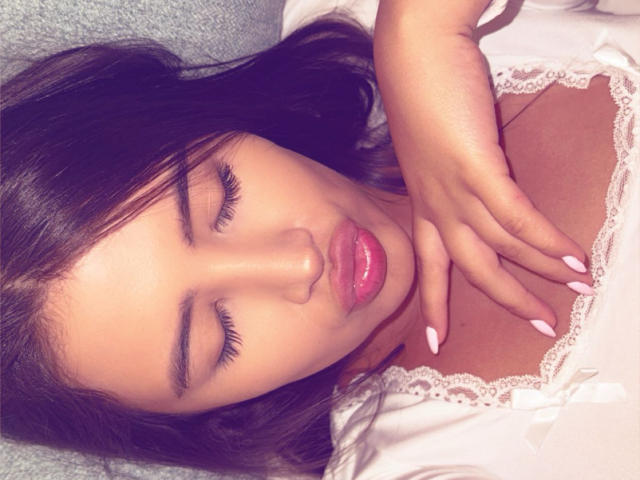 AnnaPooky with her eyes closed, dark wavy hair, glossy pink lips, white lace-trimmed top, and manicured nails resting on her chest. Her hand is on her tits.