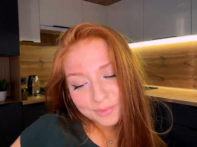 LuanneKadri has red hair, long eyelashes, and a small silver necklace. She's smiling with her eyes closed, wearing a black top, in a modern kitchen.