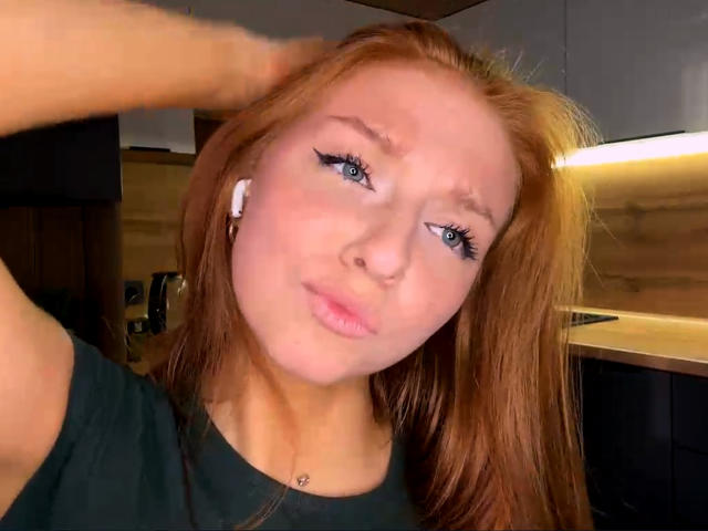 LuanneKadri with red hair, blue eyes, black eyeliner, wearing earbuds, black shirt, hand in hair, pouty lips, slight frown, in a kitchen with wooden and dark cabinets.