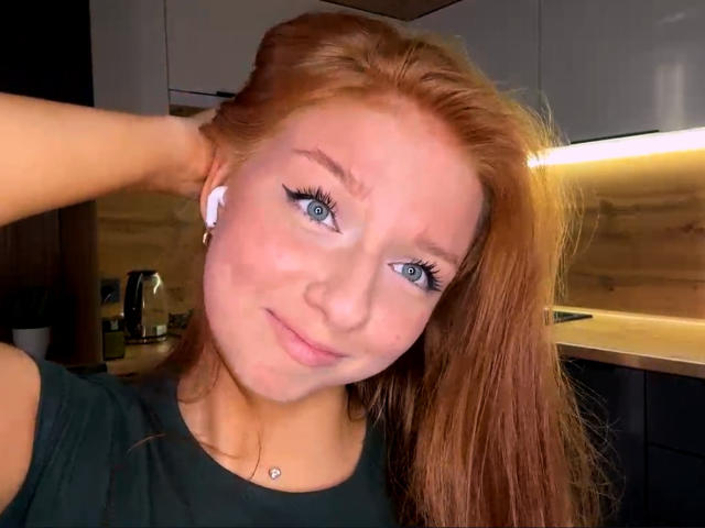 LuanneKadri has red hair, blue eyes, and a cute face with a small smile. She's wearing a green shirt, white earbuds, and a necklace with a small pendant.