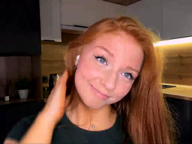 Luanne Kadri, con cabello rojo, ojos azules y un pequeño piercing en la nariz, sonríe suavemente mientras una mano le toca la cabeza. Lleva una camisa negra y está en una cocina moderna..