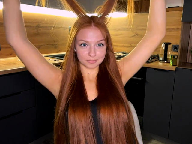 LuanneKadri has long red hair, arms raised, wearing a black sleeveless top, in a modern kitchen with wooden and dark cabinets, looking directly at the camera with a slight smile.