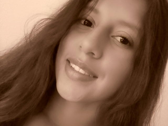 ArenaPeeachs has a smiling face with long, wavy brown hair, light skin, and small, cute nose. Her eyes are slightly squinted, and she's wearing a subtle, natural makeup.