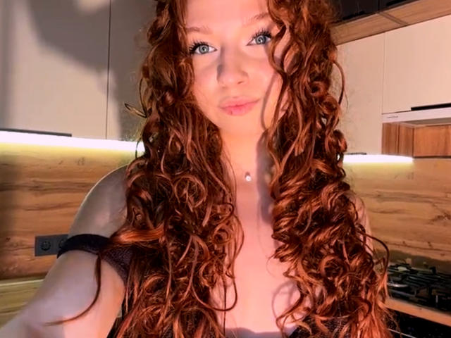 LuanneKadri with long, curly red hair, green eyes, and pale skin, wearing a black strap top, has small tits and a delicate necklace in a brightly lit kitchen.