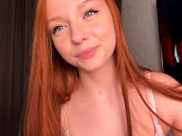 LuanneKadri has red hair, blue eyes, and a small silver necklace. She's smiling with pouty lips, wearing a pink top, and has a cute freckled face.