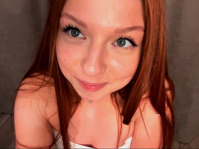 LuanneKadri with red hair, blue eyes, and a cute smile, wearing a white top, showing her small tits and a pierced nose. Her makeup includes long, dark eyelashes.