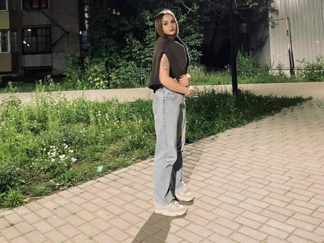 ShannanVasmadjides stands on a brick path, wearing a black top, blue jeans, and white sneakers. She has a tattoo on her right arm and a sexy, confident look.