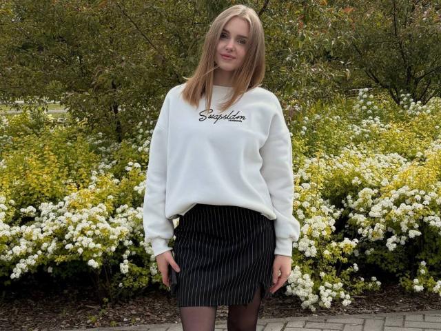 ShannanVasmadjides with blonde hair, white sweatshirt, black pinstriped skirt, and black tights, standing in front of white flowers and greenery. She's looking at the camera with a slight smile.