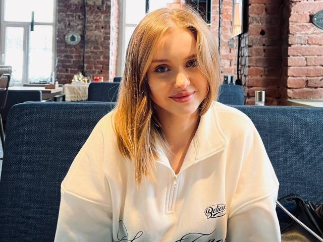 ShannanVasmadjides has blonde hair, fair skin, and is wearing a white polo shirt, sitting on a blue chair, smiling with pink lips. Her tits are small and she's cute.