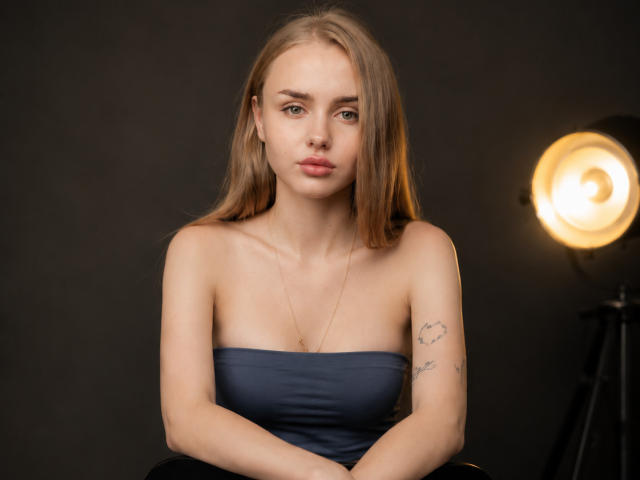 ShannanVasmadjides, blonde, strapless blue dress, small tits, light skin, tattoo on right arm, serious expression, soft light from right, sitting, dark background, subtle makeup, necklace.