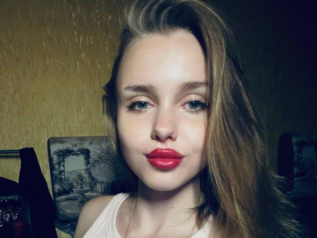 ShannanVasmadjides has pale skin, green eyes, and bright red lipstick. She's wearing a white top with long, straight brown hair covering one side of her face.