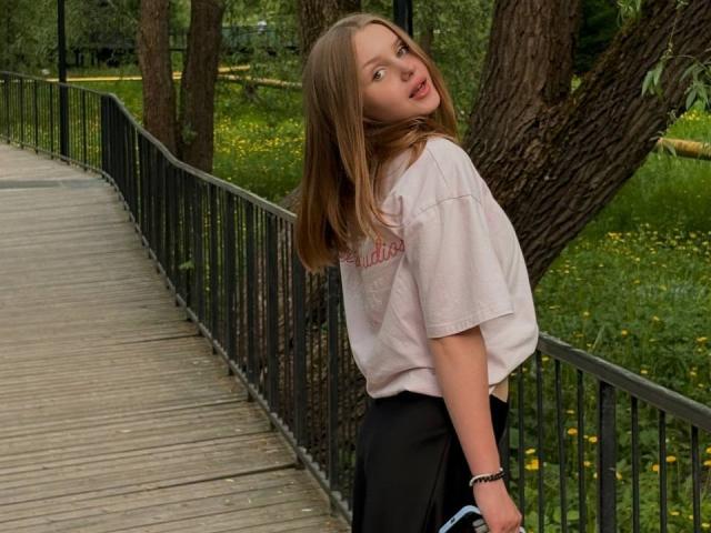 ShannanVasmadjides with light brown hair, looking over her shoulder, wearing a loose pink shirt and black pants, standing on a wooden bridge, holding a blue bag.