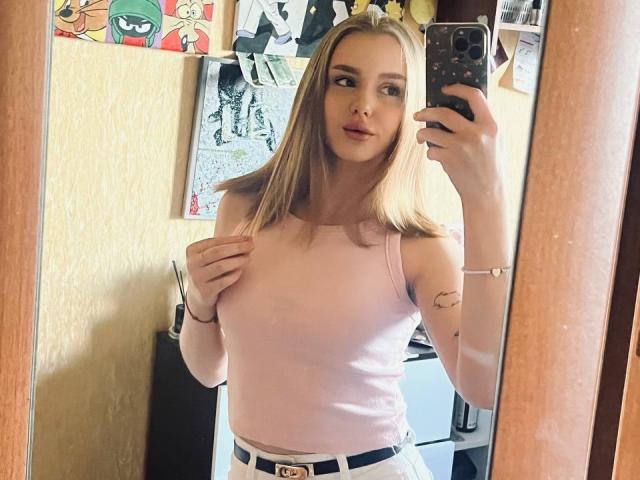 ShannanVasmadjides with long blonde hair, pink tank top, white pants, holding phone in mirror selfie. Her tits are small, she's looking at camera with a slight smile.