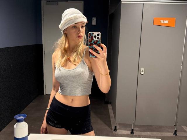 OliviaRenly with blonde hair, white beanie, gray crop top, black shorts, holding phone in mirror, visible boobs, small ass, locker room, bottle on counter, reflective surface.