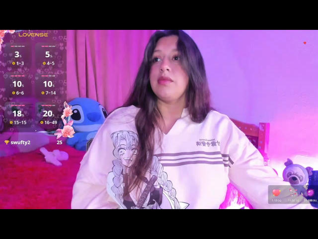 ArenaPeeachs has long brown hair, wearing a white long-sleeve shirt with a graphic, sitting in a room with a blue stuffed toy and pink lighting.