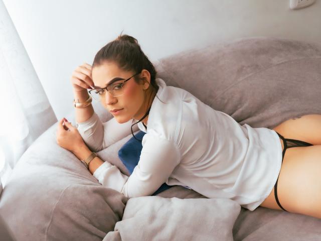 Alice Coolenk, allongée sur un canapé gris, portant des lunettes, une chemise blanche, un string noir et une cravate bleue, avec sa main sur sa tête, montrant ses seins et ses fesses..