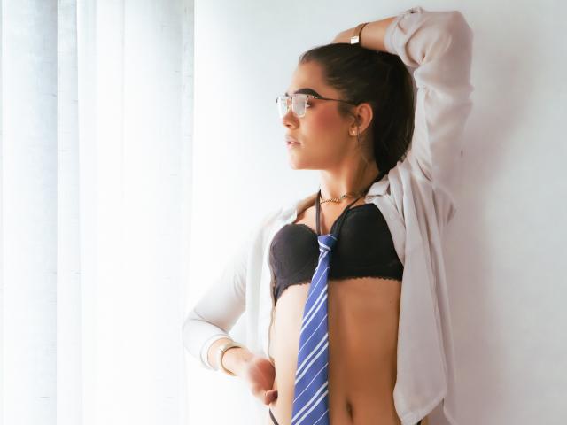 AliceCoolenk, avec des lunettes, chemise blanche ouverte, soutien-gorge noir, cravate bleue à rayures, main dans les cheveux, debout près d'une fenêtre, regardant sur le côté, peau bronzée, cheveux bruns attachés en queue de cheval..