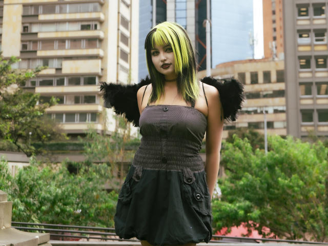 AngelBlakee with green and black hair, dark lipstick, black strapless dress, fluffy black shoulder accessories, standing in urban area, hands in pockets, looking forward, medium shot.