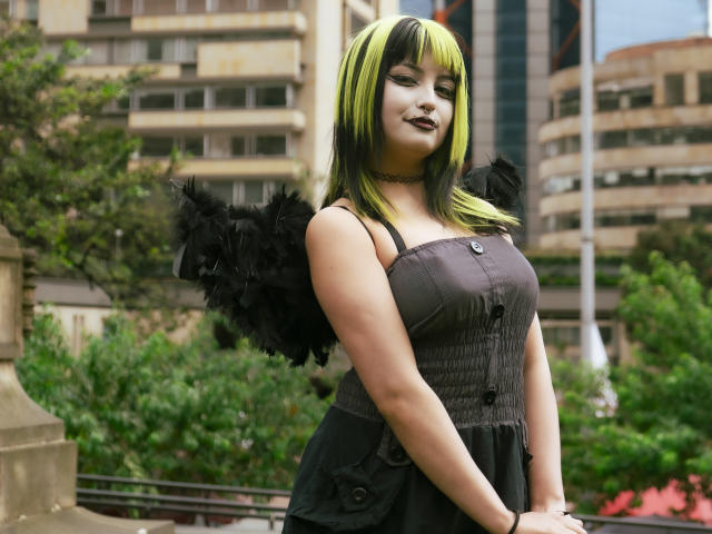 AngelBlakee with black feather wings, green and black hair, dark lipstick, choker, and black dress, showing her big ass in a city park, standing with a confident smile.