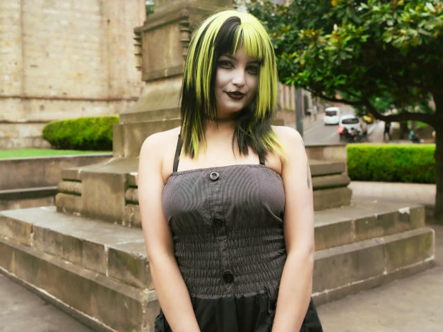 AngelBlakee, com cabelo preto e verde, batom escuro, vestido com alças cinzas, mostrando os seios, em pé em frente a um monumento de pedra, com as mãos ao lado do corpo, olhando para a câmera..