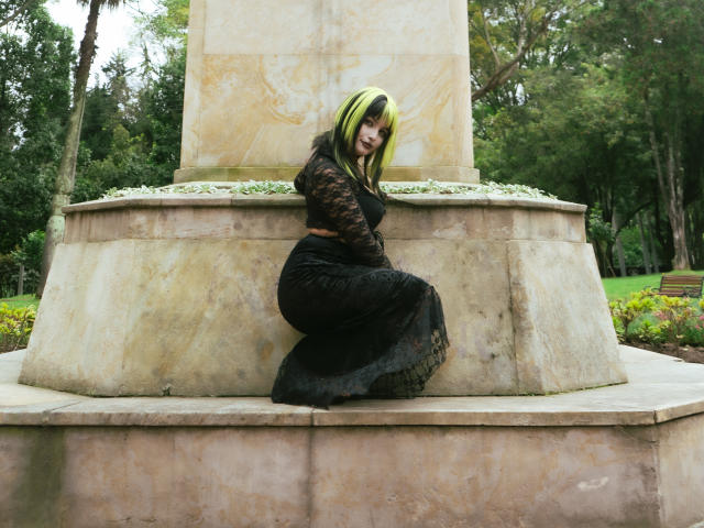 AngelBlakee with green and black hair, wearing a black lace dress, crouching in front of a stone fountain, showing her ass and looking over her shoulder.
