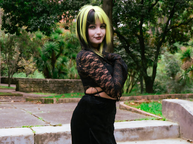 AngelBlakee, avec des cheveux verts et noirs, des manches en dentelle, une robe noire, les bras croisés, debout sur des marches de pierre, souriante, montrant ses seins, dans une zone boisée..