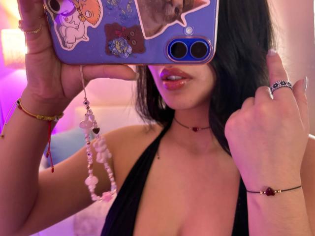 EmmaBeaumont taking a selfie with her phone, showing her glossy red lips and black halter top, with her tits partially visible, wearing a bead bracelet and ring.