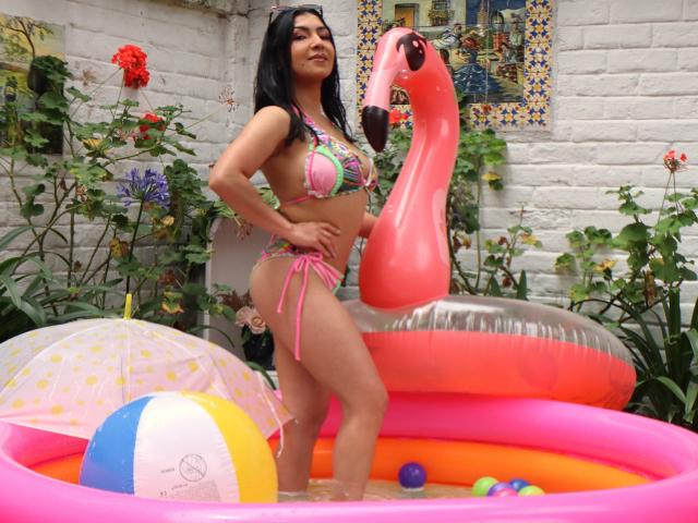 Chanel, vêtue d'un bikini coloré, se tient dans une piscine gonflable en forme de flamant rose, avec un autre flamant rose flottant, un parapluie à pois et une balle arc-en-ciel. Ses seins sont petits..