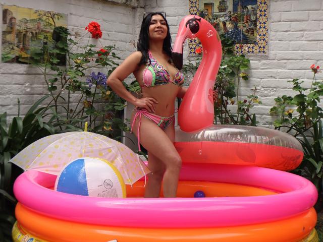 ChanelBrouw steht in einem rosa aufblasbaren Pool, trägt einen bunten Bikini, mit einem rosa Flamingo-Poolspielzeug und einem Strandball, wobei ihre Brüste und ihr Hintern sichtbar sind, und lächelt..