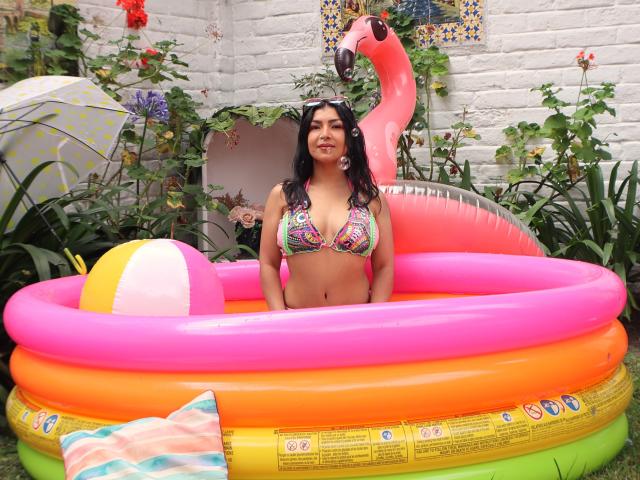 ChanelBrouw with medium tits in a colorful bikini, sitting in a pink inflatable pool, smiling, with a pink flamingo and beach ball. Her black hair is down.