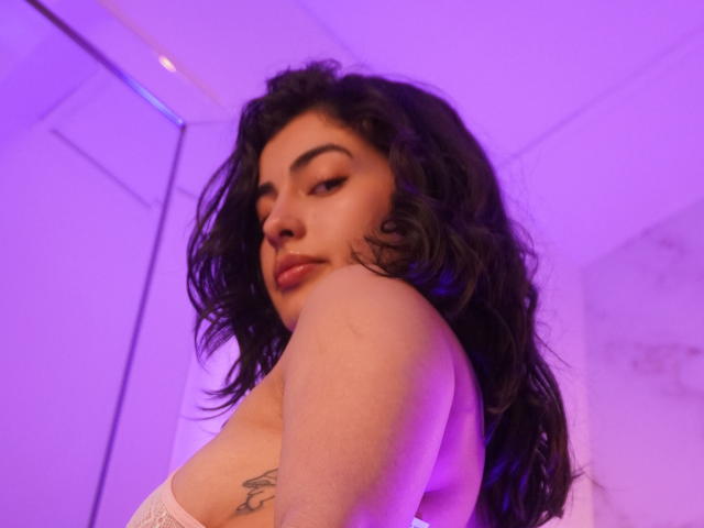 EmmaBeaumont with wavy black hair, dark eyebrows, and pinkish-purple lighting, looking over her shoulder, showing her bare shoulder and tattoo on her upper arm. Boobs.