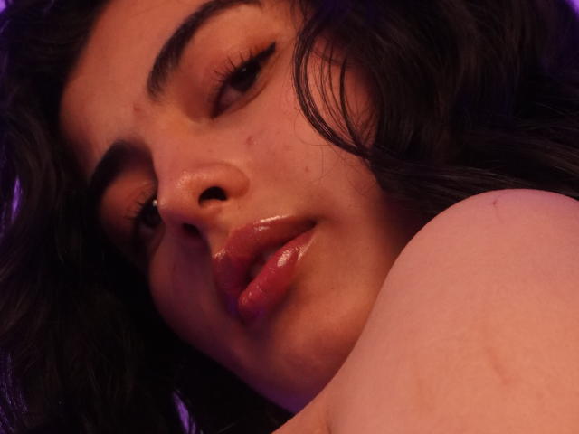 EmmaBeaumont's face with glossy pink lips, dark wavy hair, and subtle makeup, looking slightly to the side with a relaxed expression. Her bare shoulder is visible in the foreground.