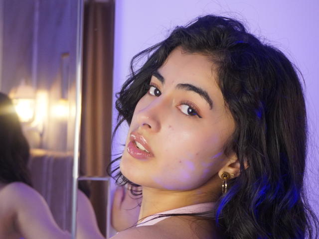 EmmaBeaumont with wavy black hair, brown eyes, and glossy pink lips, wearing a pink halter top, looking over her shoulder, showing her bare shoulders and gold hoop earrings.