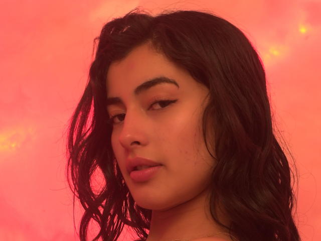 EmmaBeaumont has medium brown skin, wavy black hair, and subtle makeup with winged eyeliner. She's looking down with a slight pout, against a pink, glowing background.