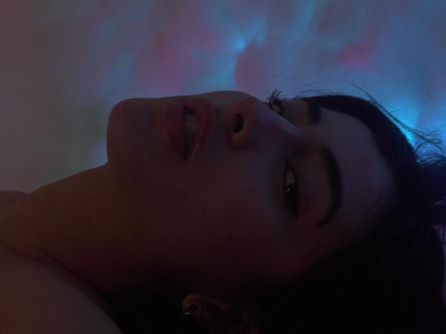 EmmaBeaumont's face, dark hair, parted lips, slightly open eyes, subtle makeup, dim blue and pink light, looking up, soft shadows, nose slightly upturned, dark eyebrows, relaxed expression.