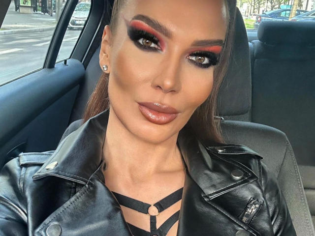 MistressKarolina-hot with glossy brown lips, bold red and black eye makeup, strappy black top, leather jacket, and earrings, sitting in a car, looking at the camera.