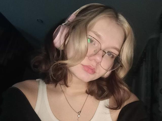 DorcasHarmen with blonde and brown wavy hair, glasses, pink headphones, pouty pink lips, white tank top, black off-shoulder top, necklace, green eye reflection, dimly lit.