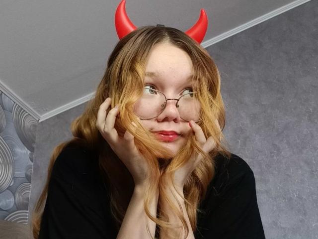 DorcasHarmen with red devil horns, glasses, and wavy blonde hair, hands on cheeks, black top, red lipstick, looking to the side, slightly smiling, in a room.