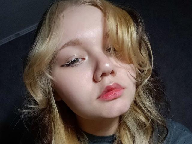 DorcasHarmen's face with blonde, wavy hair covering one eye, light skin, small nose, pink lips, subtle eyeliner, and a slightly upturned, relaxed expression. Her nose is small.