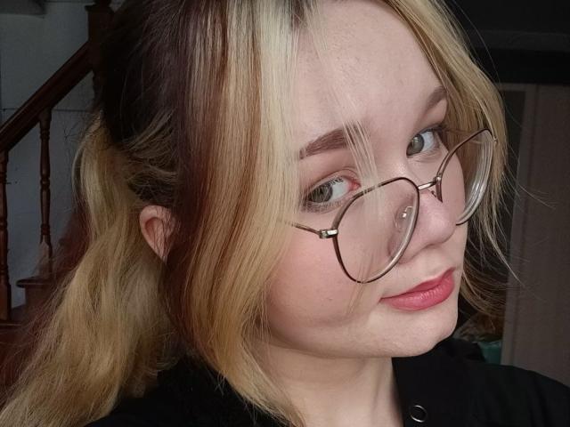 DorcasHarmen has blonde and brown hair in a ponytail, wearing glasses with a nose piercing, and has pink lips. She's looking slightly to the side with a subtle smile.