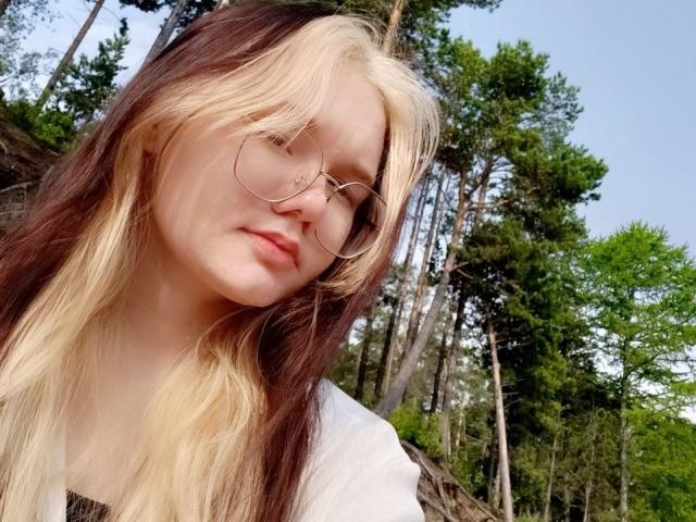 DorcasHarmen has long, brown-and-blond hair, wearing round glasses and a white shirt. She's looking down with a slight, relaxed smile. Forest in background.