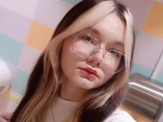 DorcasHarmen with blonde and brown hair, wearing glasses, and a white top, has a cute face and pink lips, looking at the camera with a slight pout.