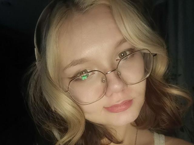 DorcasHarmen with blonde and brown wavy hair, wearing glasses, has a small nose piercing, and a slight smile, showing her tits and green reflection in her glasses.