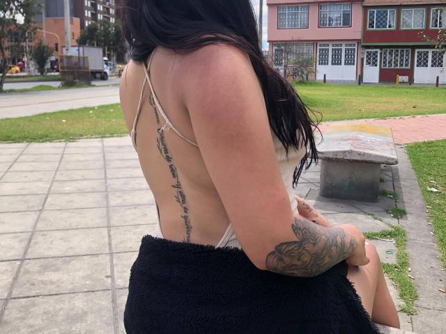 DulceRosier with long black hair, back to camera, wearing a string top, showing a tattoo down her side and a black towel covering her ass. She has a tattooed arm.