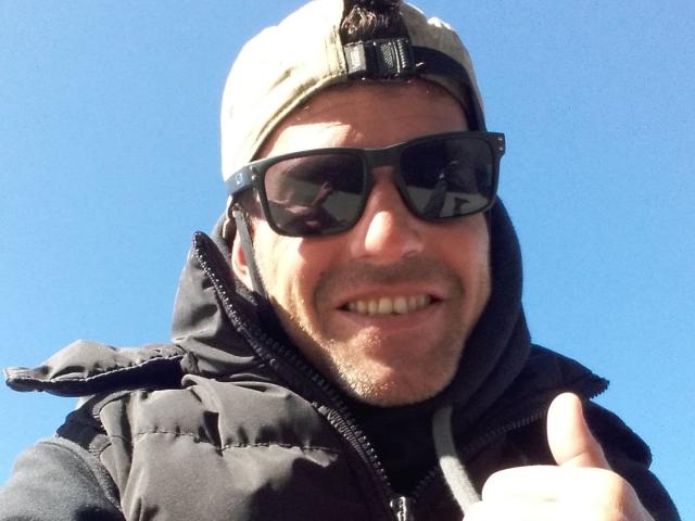 TheoCool with a beard, black sunglasses, beige cap, and black puffy jacket, giving a thumbs-up, smiling, against a clear blue sky.