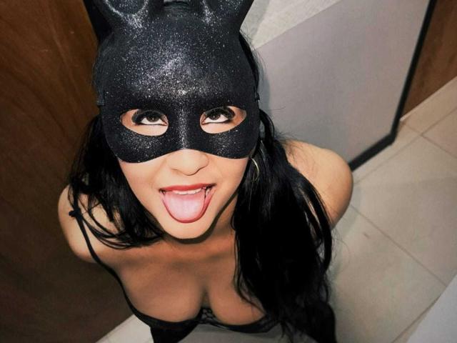 MiaRouusee com máscara preta brilhante, língua de fora, cabelo preto, seios grandes, vestindo lingerie preta, olhando para cima, porta de madeira à esquerda, chão de azulejos brancos à direita..