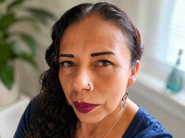 LaylaMilf tem cabelos escuros e ondulados, um piercing no nariz, batom roxo e olhos verdes. Ela está vestindo uma camisa azul com brincos rosa, olhando diretamente para a câmera..