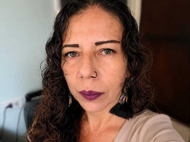 LaylaMilf has curly brown hair, purple lipstick, a nose ring, and black tassel earrings. Her green eyes are slightly squinted, and she's wearing a beige top.