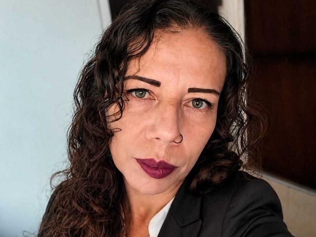 LaylaMilf with curly dark brown hair, green eyes, thick eyebrows, purple lipstick, nose ring, and wearing a black suit, looking directly at the camera with a serious expression.